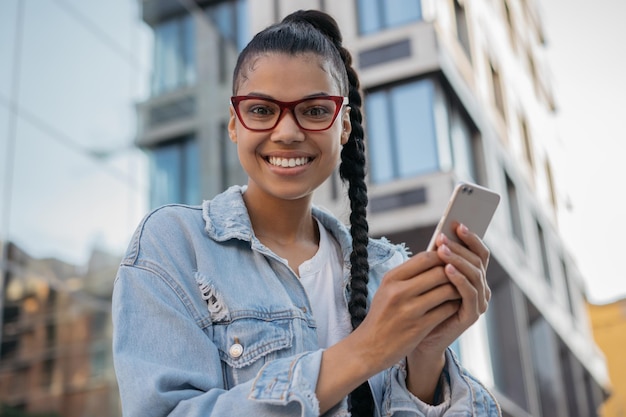 Uśmiechnięta nastolatka hipster nosząca stylowe okulary, korzystająca z aplikacji mobilnej robiąca zakupy online na świeżym powietrzu