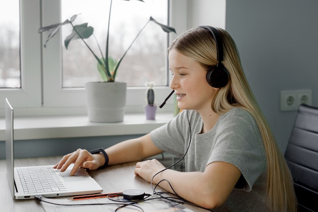 Uśmiechnięta nastolatka w słuchawkach używa swojego laptopa mając lekcję online w domu