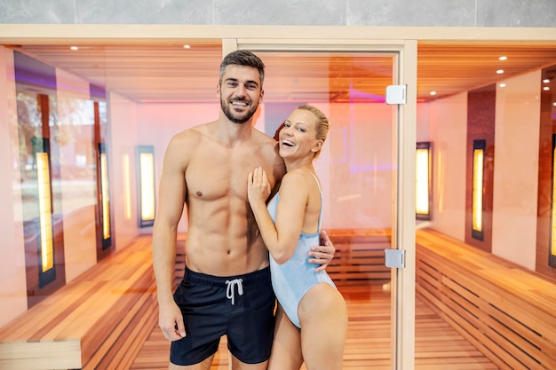 Uśmiechnięta para w strojach kąpielowych przytula się i stoi przed sauną w centrum spa