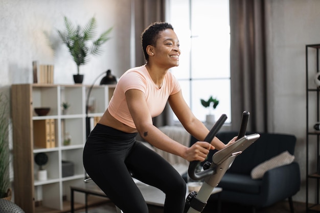 Uśmiechnięta piękna Afroamerykanka sportowa w odzieży sportowej na rowerze w domu