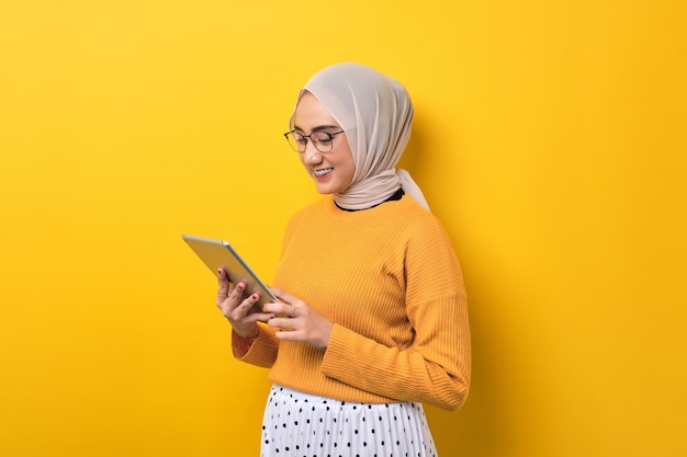 Uśmiechnięta piękna Azjatka nosząca hidżab za pomocą cyfrowego tabletu odizolowana na żółtym tle