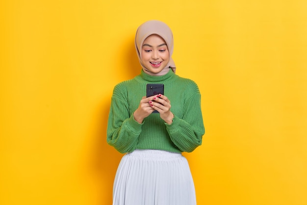 Uśmiechnięta piękna Azjatka w zielonym swetrze korzystająca z telefonu komórkowego otrzymała dobre wieści w mediach społecznościowych izolowanych na żółtym tle