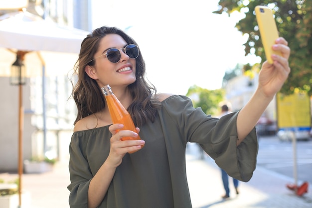 Uśmiechnięta piękna brunetka kobieta spaceru na świeżym powietrzu ulicą, weź selfie przez telefon komórkowy, pijąc sok.