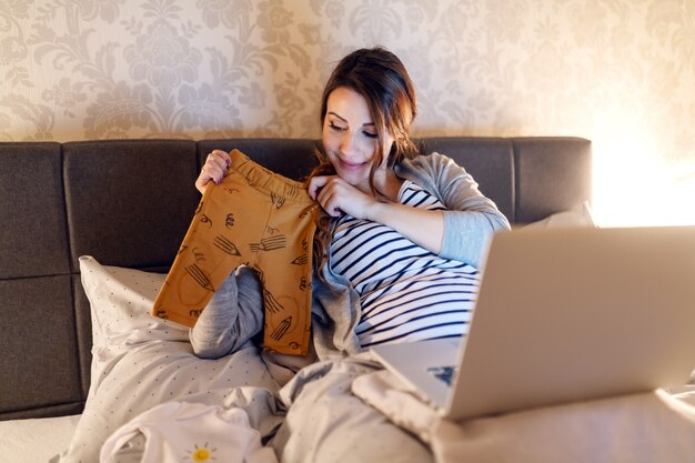 Uśmiechnięta piękna brunetka o rozmowie wideo przez laptop. Kobieta pokazująca ubrania dla dzieci, które kupiła. Porą wieczorową.