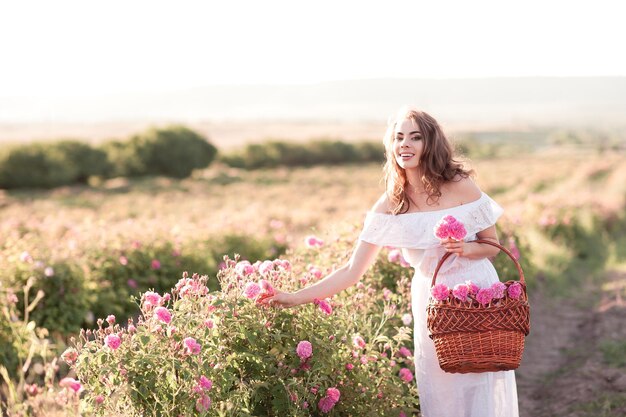 Uśmiechnięta piękna dziewczyna ubrana w białą sukienkę trzymająca kosz z różami na zewnątrz