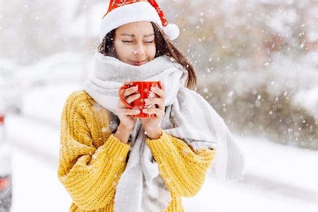 Uśmiechnięta piękna dziewczyna w kapeluszu Santa stoi na zewnątrz w śniegu. Wesołych Świąt w tle