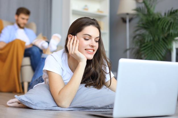 Uśmiechnięta piękna kobieta za pomocą laptopa z zamazanym mężczyzną w tle w domu.