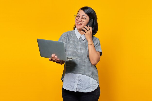 Uśmiechnięta Piękna Młoda Kobieta Rozmawiająca Na Smartfonie I Patrząca Na Ekran Laptopa Na żółtym Tle