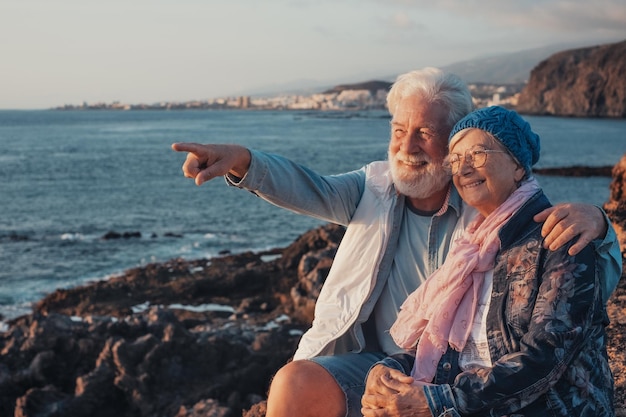 Uśmiechnięta piękna starsza para siedzi na skałach na morzu, ciesząc się światłem zachodu słońca
