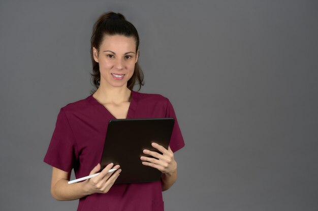Uśmiechnięta pielęgniarka w fioletowym mundurze patrzy na kamerę z raportem cyfrowego tabletu na szarym tle