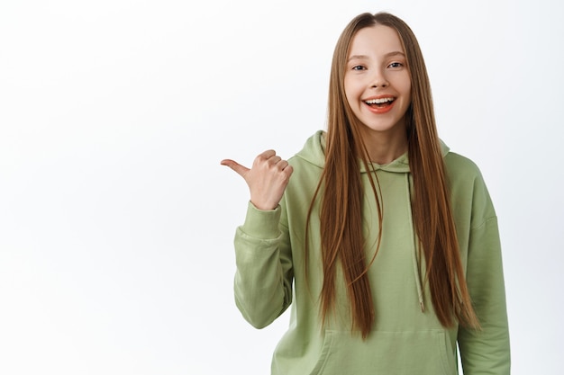 Uśmiechnięta pozytywna dziewczyna z długimi włosami, wyglądająca na szczęśliwą, wskazująca na bok, wskazująca drogę, stojąca w bluzie z kapturem na tle białej ściany.