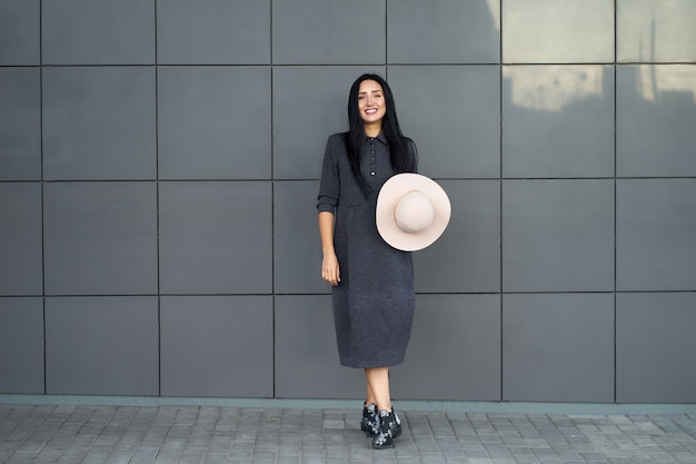 Uśmiechnięta pozytywna ładna młoda kobieta jest ubranym młodzieżową szarą obszerną suknię i modne buty trzyma modnego kapelusz. Model w modnym stroju pozowanie na szarej ścianie miejskiej. Portret na zewnątrz.