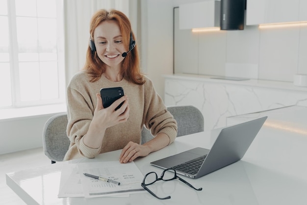 Uśmiechnięta pracownica biurowa w zestawie słuchawkowym za pomocą smartfona siedząc w miejscu pracy z laptopem