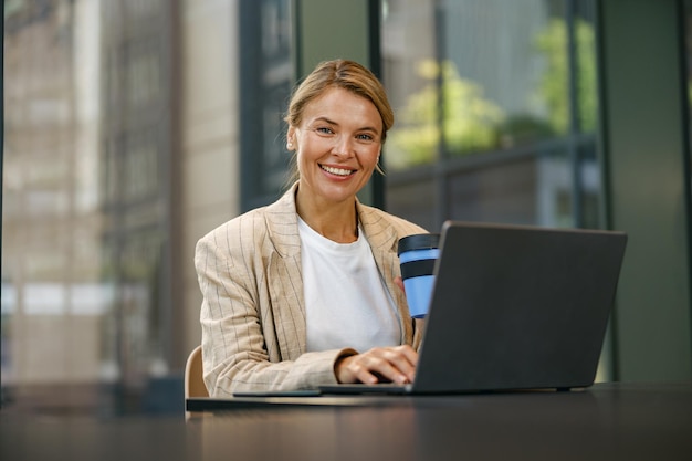 Uśmiechnięta przedsiębiorczyni pije kawę, pracuje na laptopie na tarasie biura i patrzy na kamerę.
