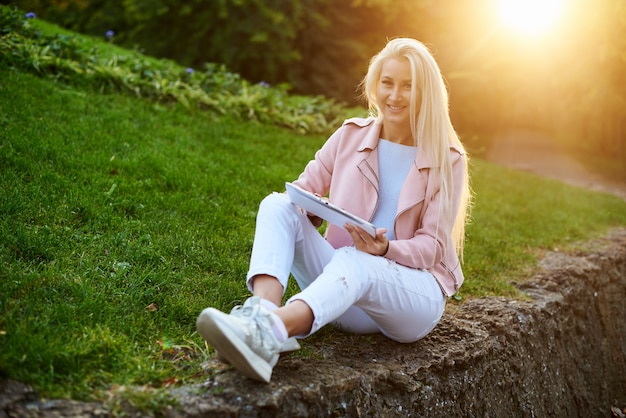 Uśmiechnięta przystojna kobieta słucha relaksującą muzykę W hełmofonach z jej telefonem komórkowym lub pastylką przy parkiem. student słucha wykładu online i przygotowuje się do egzaminów.