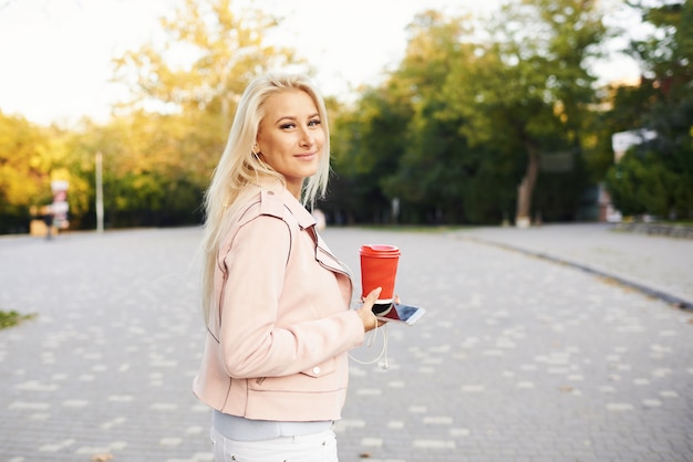 Uśmiechnięta Przystojna Kobieta Słucha Relaksującą Muzykę W Hełmofonach Z Jej Telefonem Komórkowym Lub Pastylką Przy Parkiem. Student Słucha Wykładu Online I Przygotowuje Się Do Egzaminów.