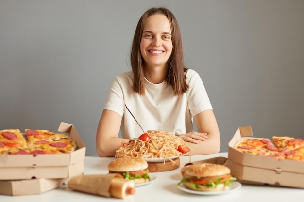 Uśmiechnięta radosna młoda kaukaska kobieta o brązowych włosach ubrana w białą koszulkę siedząca przy stole wśród fast foodów odizolowanych na szarym tle lubi jeść niezdrowe potrawy