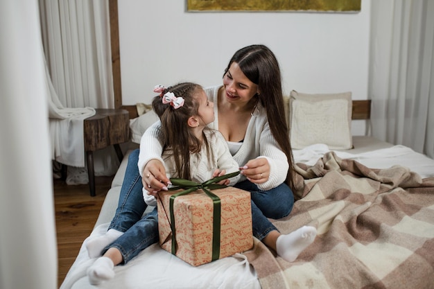 Uśmiechnięta rodzicielka daje swojej córeczce pudełko z prezentami, gratuluje mu Bożego Narodzenia, siedząc na łóżku z feriami zimowymi w przytulnym domu.