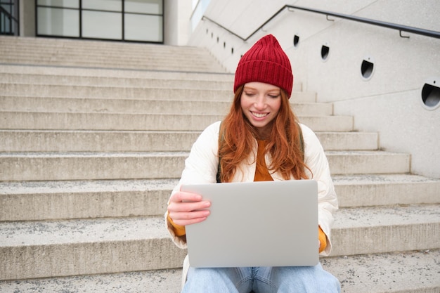 Uśmiechnięta ruda studentka siedzi na schodach na zewnątrz i korzysta z laptopa, który łączy się z publicznym Wi-Fi w mieście