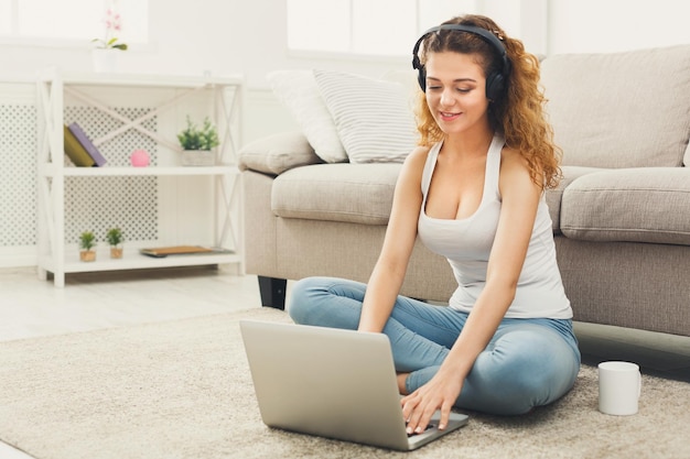 Uśmiechnięta ruda studentka używająca laptopa do nauki lub pracy, siedząca na dywanie na podłodze, słuchająca muzyki, kopiująca przestrzeń