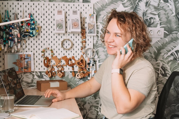 Uśmiechnięta skoncentrowana biznesowa kobieta rozmawia smartfon pracujący na laptopie w warsztacie Akcesoria do opieki zdrowotnej dla niemowląt silikonowe eko grzechotki gryzaki smoczki wiszące na białej ścianie Domowe biuro