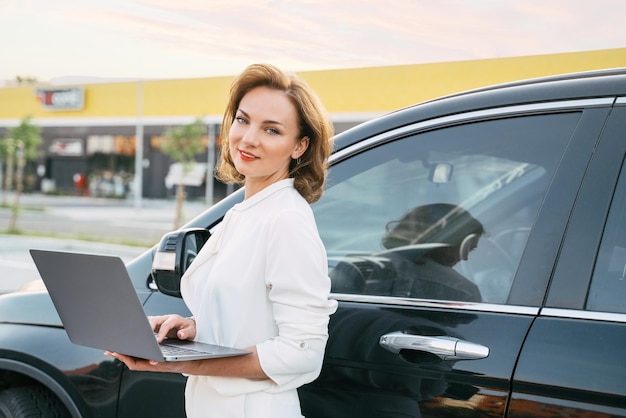 Uśmiechnięta sprzedawczyni w średnim wieku za pomocą laptopa stojącego w pobliżu nowego samochodu. Dealerzy, koncepcja wynajmu samochodów