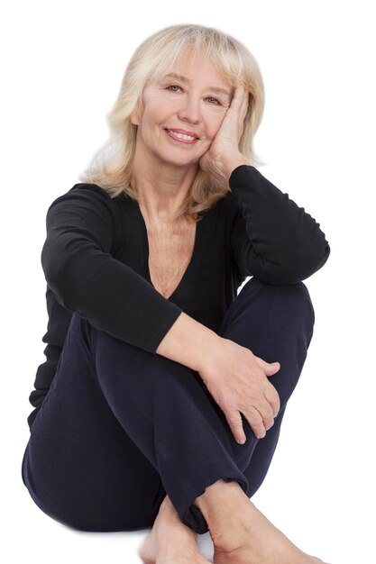 Zdjęcie uśmiechnięta starsza blondynka piękna dama w czarnym swetrze i niebieskich spodniach siedzi wdzięcznie na podłodze działalność piękno i zdrowie w dorosłości izolowana na białym tle pionowa