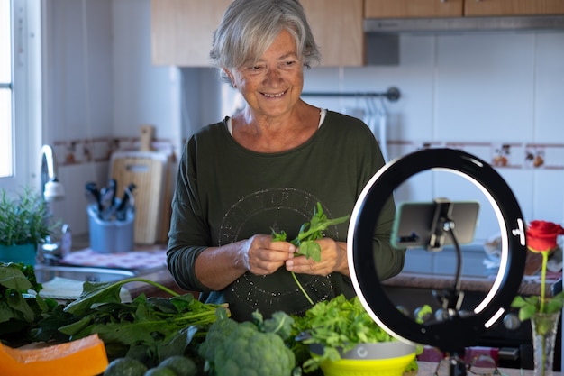 Uśmiechnięta starsza kobieta, która transmituje online, śledzi lekcje gotowania wegetariańskiego. Domowy stół kuchenny pełen zdrowych warzyw