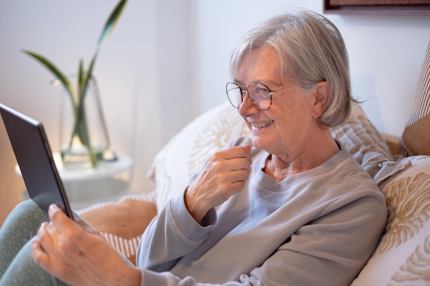 Uśmiechnięta starsza kobieta w sypialni w domu lub pokoju hotelowym, wyglądająca na cyfrowy tablet, ciesząca się technologią i społecznością