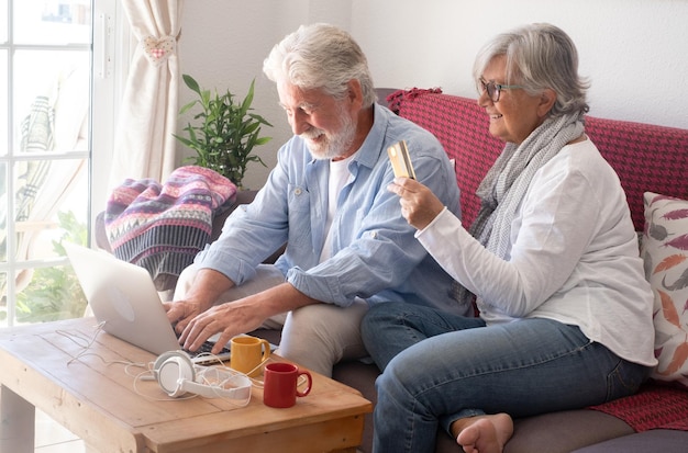 Uśmiechnięta starsza para robi zakupy online przez laptopa siedząc na kanapie w domu Starzy atrakcyjni siwowłosi ludzie płacą kartą kredytową Bank Konsumpcjonizm koncepcja