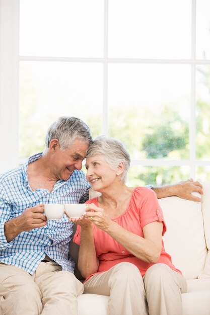 Uśmiechnięta starsza para wznosi toast z kubkami na kanapie