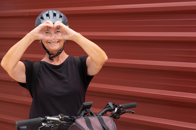 Uśmiechnięta starsza rowerzystka w miejskim mieście z elektrycznym rowerem w pobliżu czerwonego metalowego panelu Zdrowy styl życia dla emerytów i koncepcja zrównoważonej mobilności
