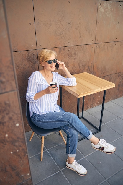 Uśmiechnięta stylowa blond kobieta w okularach przeciwsłonecznych rozmawia przez telefon komórkowy w ulicznej kawiarni