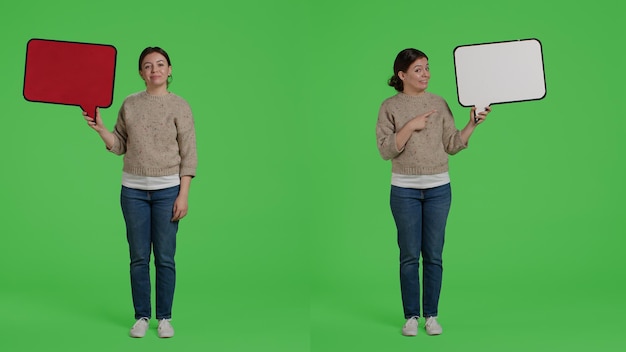 Uśmiechnięta szczęśliwa dziewczyna trzyma kartonową bańkę mowy, aby stworzyć reklamę w studio, pokazując kartonową ikonę przed kamerą. Wesoła modelka z deską stojącą na tle greenscreen.