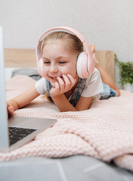Uśmiechnięta szczęśliwa dziewczynka dziecko za pomocą laptopa w bezprzewodowych słuchawkach w łóżku w domu.