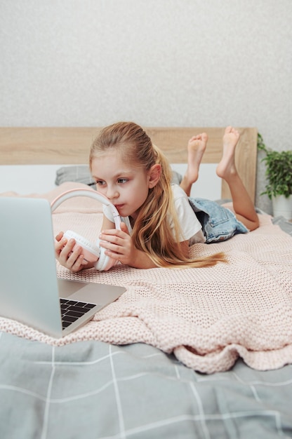 Uśmiechnięta szczęśliwa dziewczynka dziecko za pomocą laptopa w bezprzewodowych słuchawkach w łóżku w domu.