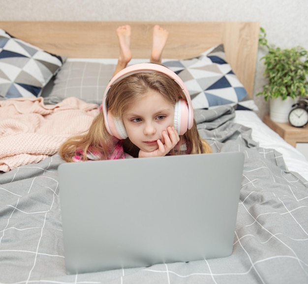 Uśmiechnięta szczęśliwa dziewczynka używa laptopa w bezprzewodowych słuchawkach w łóżku w domu