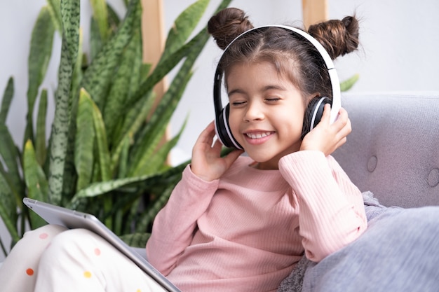 Uśmiechnięta szczęśliwa dziewczynka używa tabletu w bezprzewodowych słuchawkach na kanapie w domu