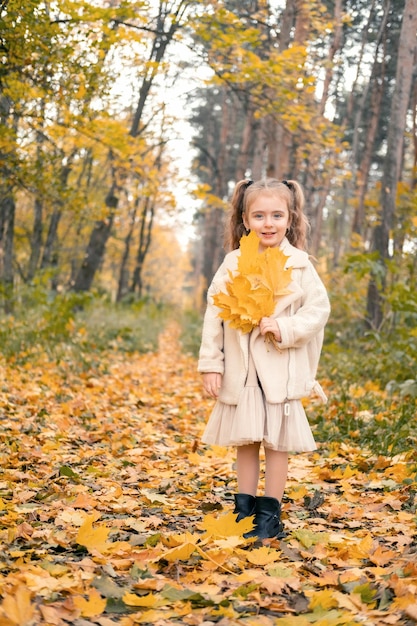 uśmiechnięta szczęśliwa dziewczynka w płaszczu i sukience trzymająca jesienne liście, bawiąca się w jesiennym lesie