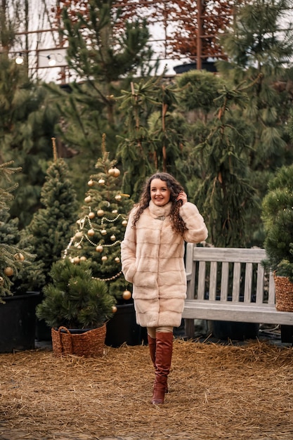 Uśmiechnięta szczęśliwa kędzierzawa kobieta w futrze krąży wśród choinek na ulicy Czekając na Boże Narodzenie i Nowy Rok