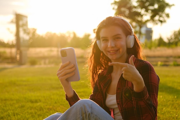 Uśmiechnięta szczęśliwa kobieta w słuchawkach wskazuje palcem na smartfon siedzący na trawie w