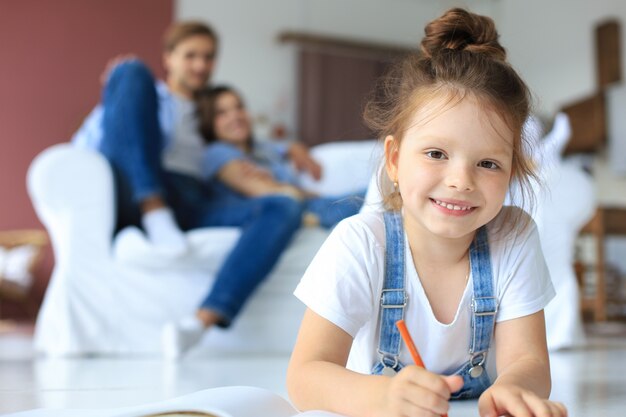 Uśmiechnięta Szczęśliwa Rodzina Siedzi Na Kanapie W Salonie I Ogląda Małą Córeczkę Rysującą W Albumie Kolorowymi Ołówkami. Wesołych Weekendów W Domu.