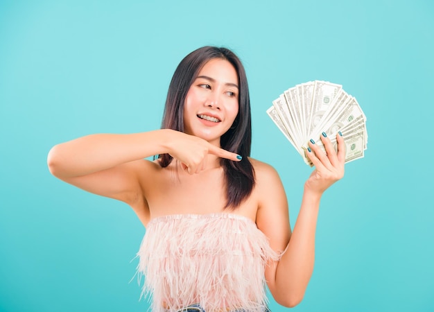 Uśmiechnięta twarz portret azjatyckiej pięknej kobiety wesoła trzyma i wskazuje pieniądze na niebieskim tle, z miejscem na kopię tekstu