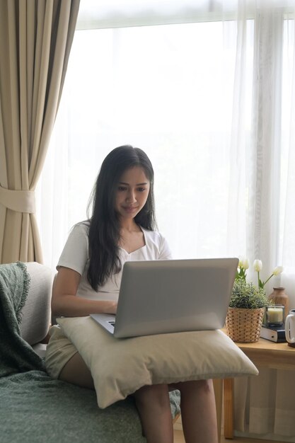 Uśmiechnięta tysiącletnia kobieta siedzi na kanapie w salonie i korzysta z laptopa