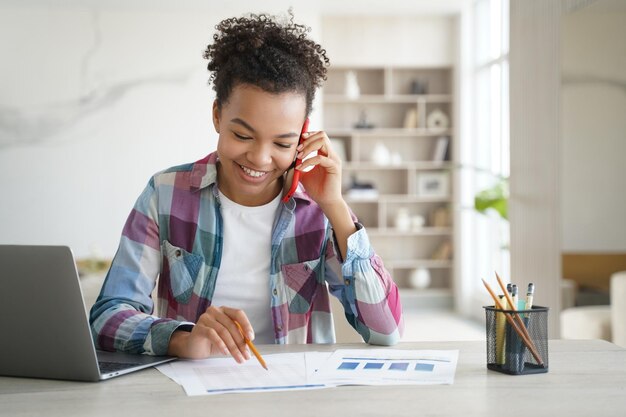 Uśmiechnięta uczennica rasy mieszanej uczy się przez telefon w domu przy biurku z laptopem