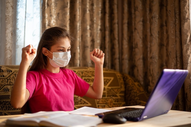 Uśmiechnięta Uczennica Studiuje W Domu W Medycznej Masce. Szczęśliwy Uczeń, Który Ma Lekcje W Domu Przez Internet, Pracuje Na Laptopie, Chętnie Poprawnie Wykonuje Zadanie