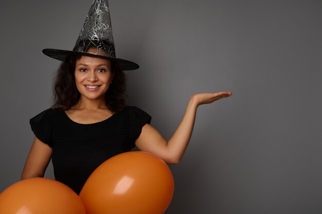 Uśmiechnięta urocza kobieta w magicznym Halloweenowym kapeluszu patrzy w kamerę i trzyma pomarańczowe balony w jednej ręce, a drugą podnosi dłoń do góry, trzymając wyimaginowaną kopię miejsca na reklamę na szarym tle. Halloween