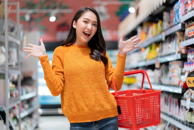 Uśmiechnięta wesoła i radosna kobieta kobieta ręka trzyma koszyk na zakupy gest ręki powitanie niespodzianka stojąca między supermarketem półka z produktem sklep spożywczy supermarket supermarket centrum handlowe