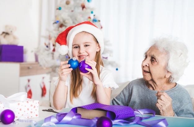 Uśmiechnięta Wnuczka W Santa Hat Siedzą Z Babcią Trzymającą Ozdobne Kulki Na Boże Narodzenie