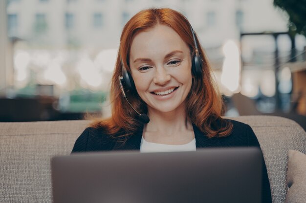 Uśmiechnięta zadowolona młoda rudowłosa kobieta nosząca zestaw słuchawkowy rozmawiająca z kolegami online na laptopie